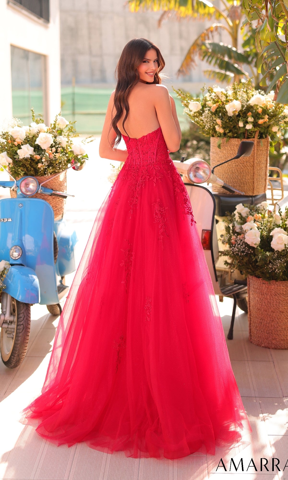 A woman in the Amarra 88950 Strapless Embroidered Prom Ball Gown stands outdoors, facing away, with blue scooters and baskets of white flowers providing a charming backdrop for her elegant red dress.